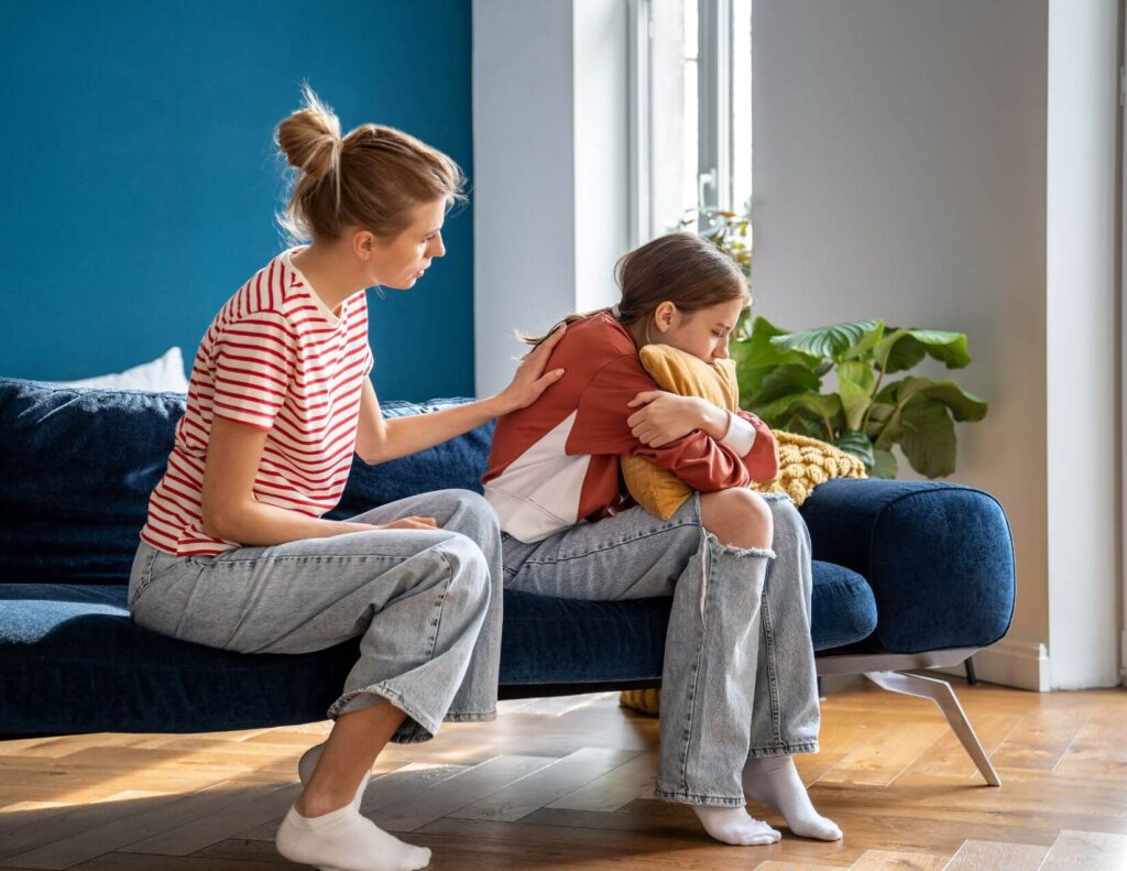 A woman comforts her teenage daughter while they sit on a couch.