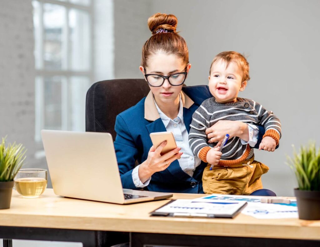 Woman holding a baby while sitting at a computer desk and looking at her mobile phone.