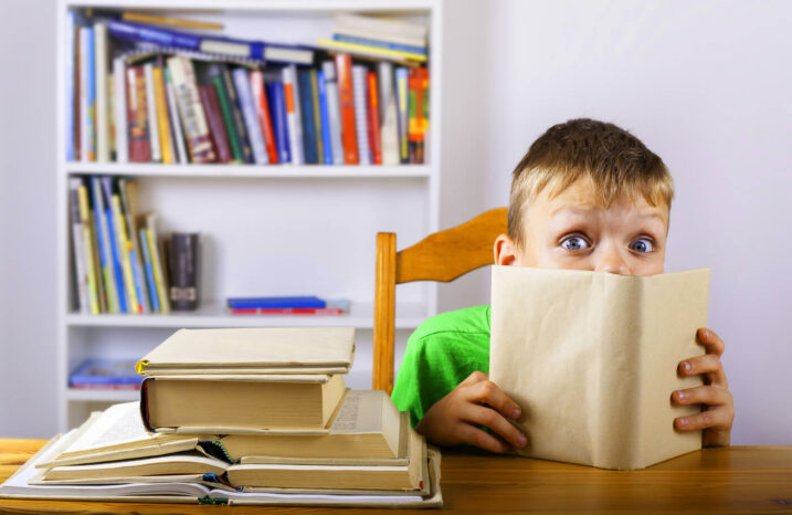 Boy sitting at a desk and peeking over the top of an open book.